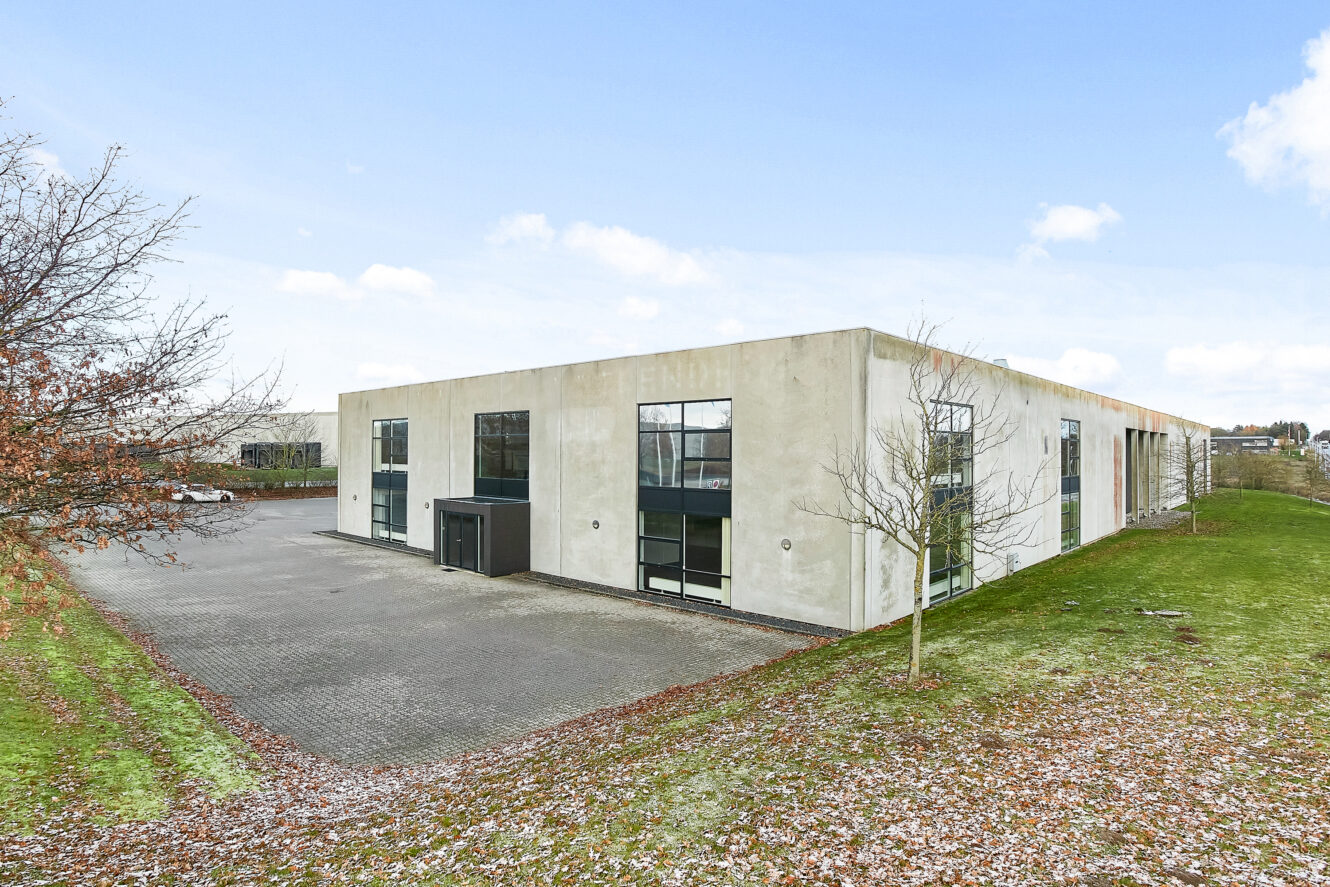 En moderne industri- eller kontorbygning i én etage med store vinduer og en asfalteret parkeringsplads, omgivet af græs og træer med nedfaldne blade.