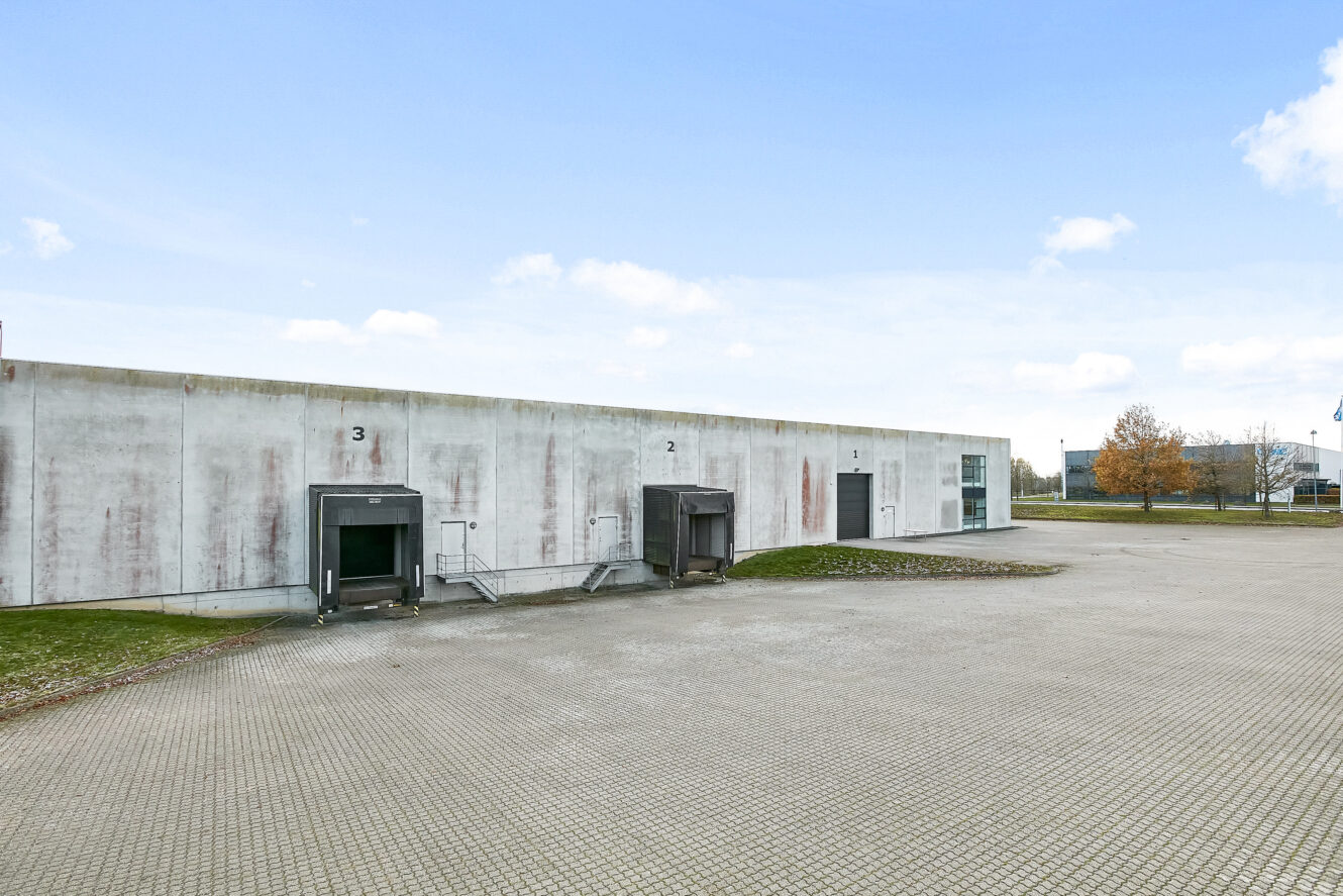 Stor hvid industribygning med tre nummererede læsseramper og et bredt asfalteret område foran; sparsomt græs og et par træer i baggrunden under en blå himmel.