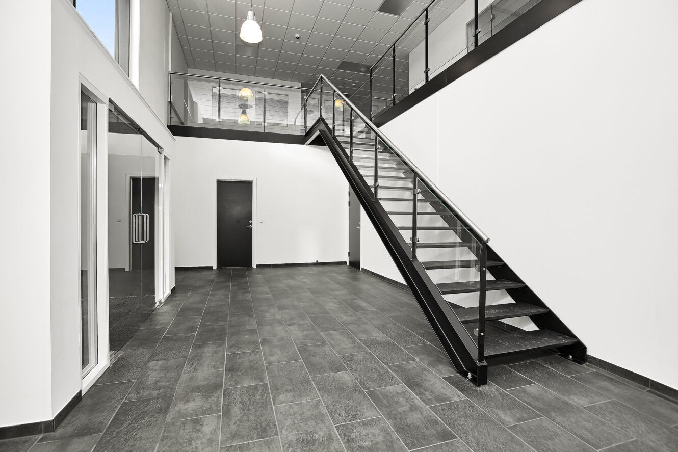 Moderne interiør med klinkegulv, kontor med glasvægge til venstre, sort dør på bagsiden og en trappe med glasgelænder, der fører til en øvre etage.