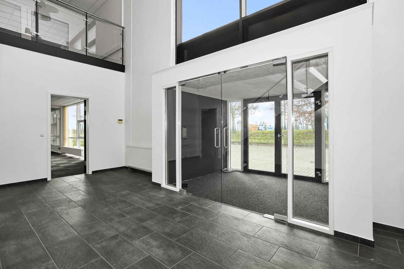Moderne kontorlobby med mørkt flisegulv, dobbelte glasdøre, hvide vægge og store vinduer, der lukker naturligt lys ind.