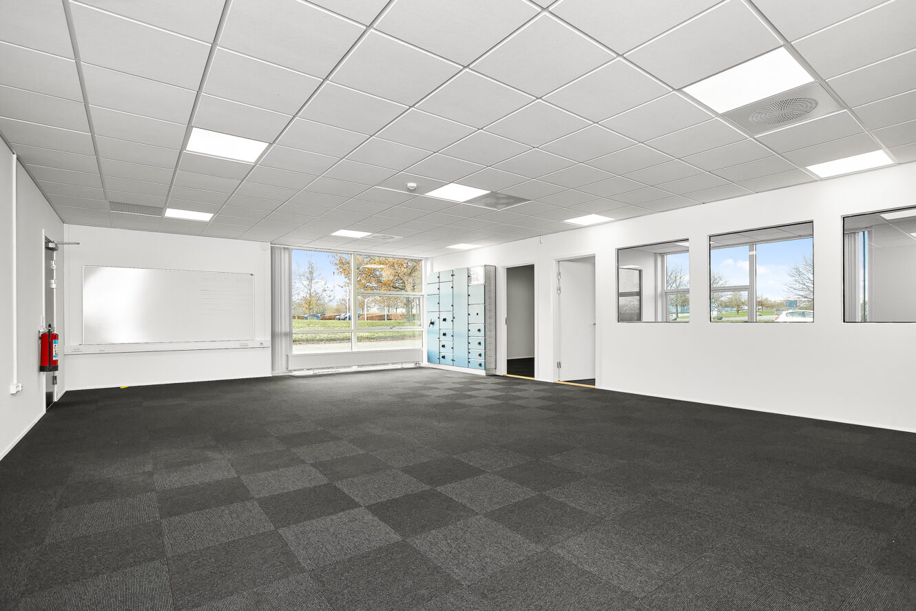 Et tomt, moderne kontorlokale med sort gulvtæppe, hvide vægge, et whiteboard, store vinduer og glasdøre, der lukker naturligt lys ind.