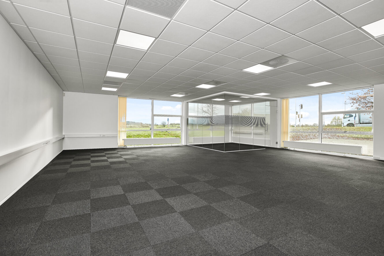 Tomt kontorlokale med ternet mørkt gulvtæppe, hvide vægge, store vinduer og et lille rum med glasvægge i hjørnet. Naturligt lys fylder rummet.