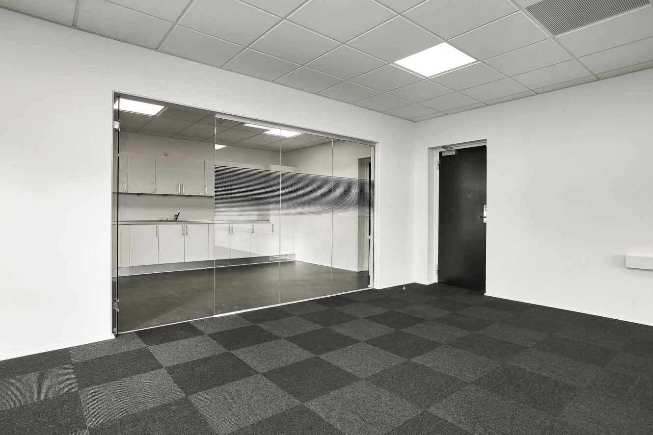 Et moderne, tomt kontorlokale med ternet tæppe, glasvæg og udsigt til et tekøkken med hvide skabe og en vask.