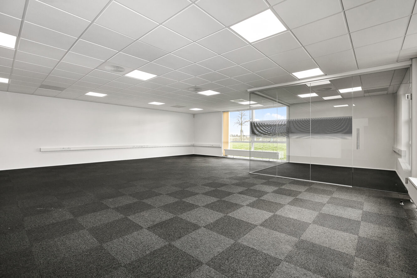 Et tomt kontorlokale med et ternet mørkt gulvtæppe, hvide vægge, et mødelokale med glasvægge og store vinduer, der lukker naturligt lys ind.