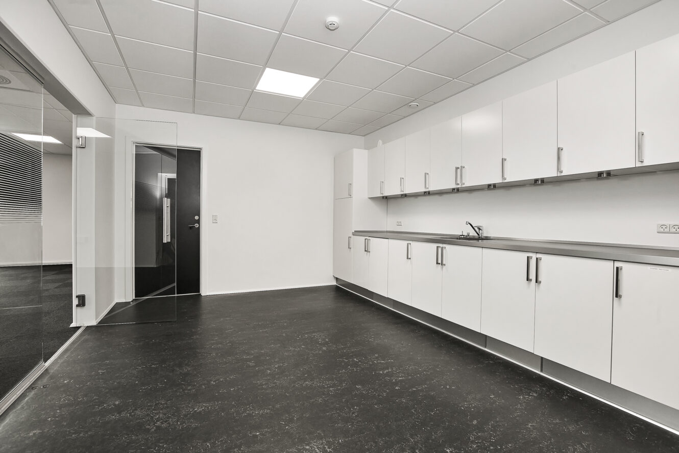 Et moderne, tomt tekøkken med hvide skabe, en sort bordplade, en vask og et sort gulv, placeret i et forretnings- eller kontorlokale.