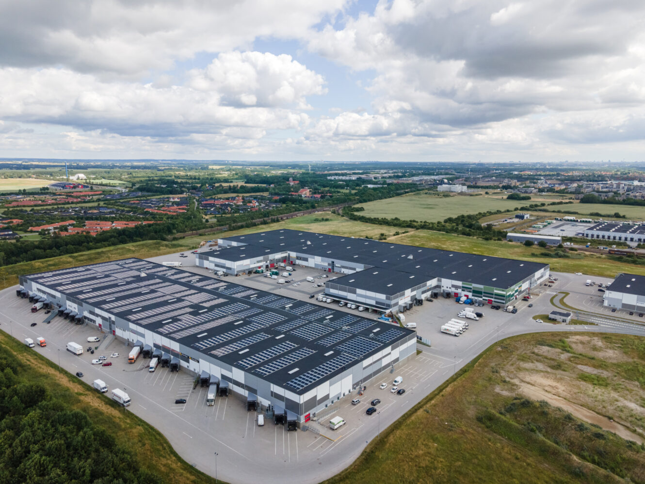 Luftfoto af et stort industrikompleks med flere lagerbygninger, parkeringspladser og flere køretøjer, omgivet af marker og spredte bygninger under en delvist overskyet himmel.