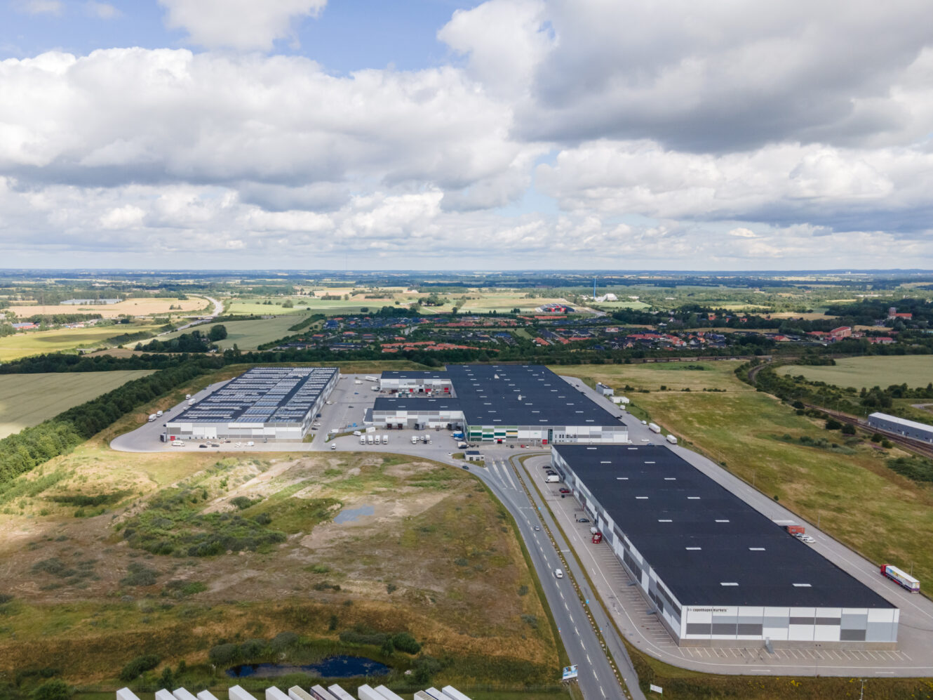 Luftfoto af et stort industrikompleks med flere lagerbygninger, omkringliggende veje og grønne marker under en delvis overskyet himmel.