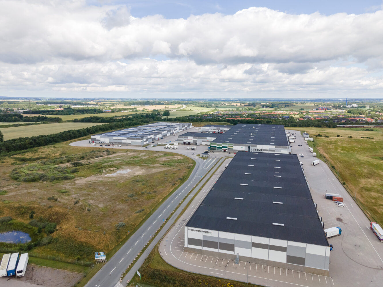 Luftfoto af et stort industrielt lagerkompleks omgivet af veje, marker og spredte lastbiler under en delvis overskyet himmel.