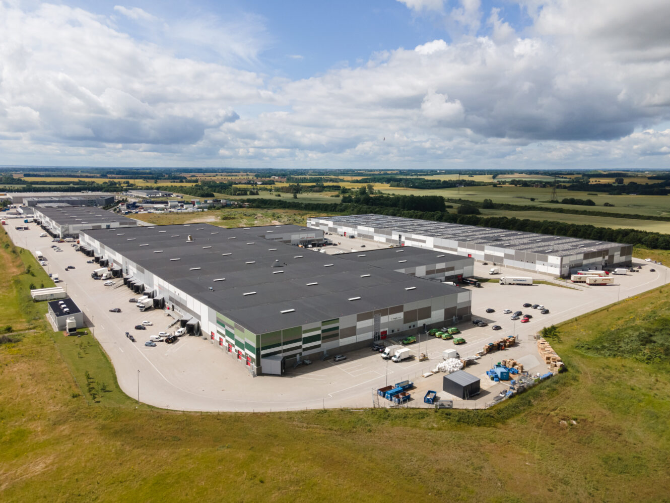 Luftfoto af et stort industrielt lagerkompleks omgivet af parkeringspladser og åbne marker under en delvis overskyet himmel.