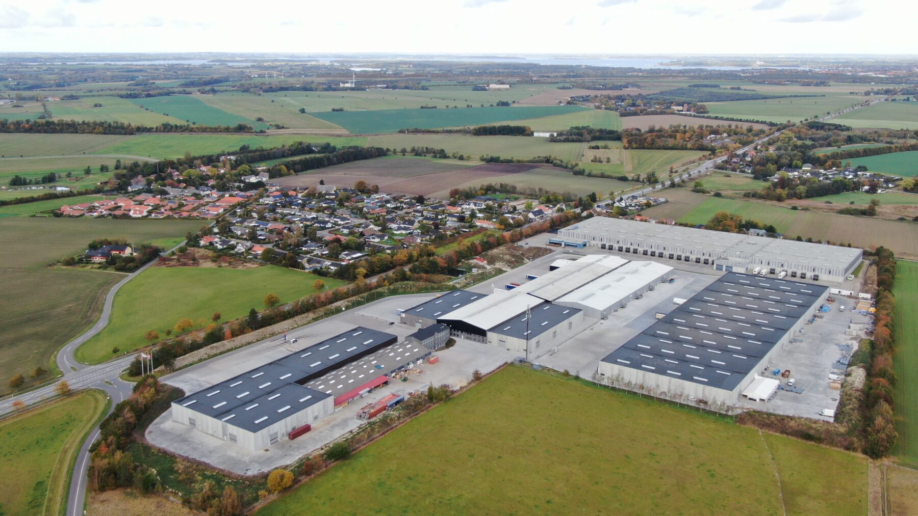Luftfoto af et stort industrianlæg med flere bygninger, marker og et nærliggende boligområde under en overskyet himmel.