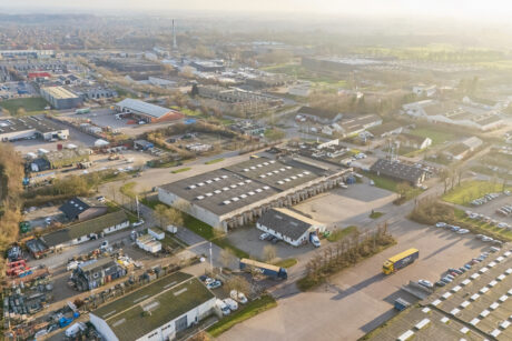 Luftfotoet viser lagerbygninger, parkerede lastbiler, biler og veje i et semi-urbant industriområde på en diset dag.