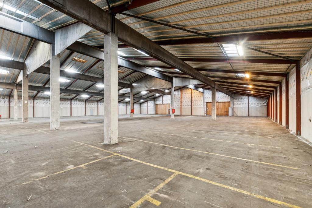 Stor tom lagerhal med højt til loftet, synlige bjælker, betongulve, markerede parkeringslinjer; oplyst af lysstofrør og ovenlysvinduer.