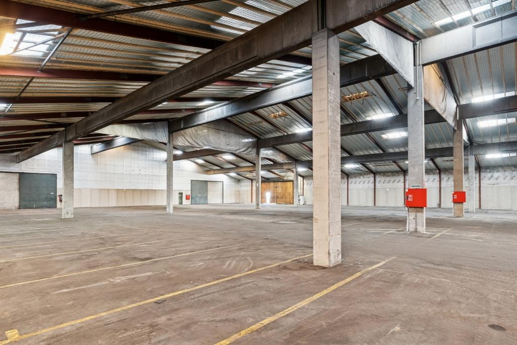 Interiør i et tomt industrilager med betongulv, metalbjælker, højt til loftet og brandslangeskabe på søjler.
