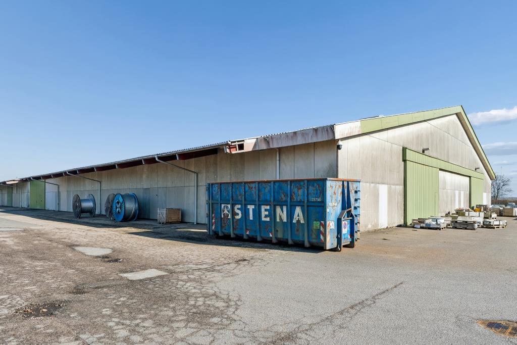 Stort industrilager med grønt tag, blå ASTENA-container foran, kabelruller og paller på revnet fortov under klar himmel.