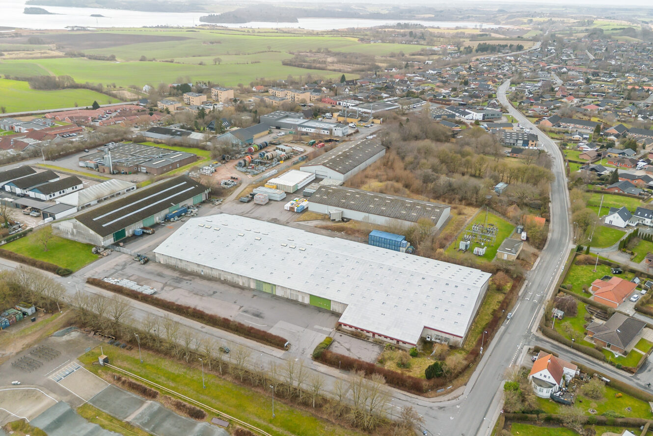 Luftfoto af et industriområde med lagerbygninger, veje, beboelsesejendomme, marker og vand i et semi-landligt landskab.