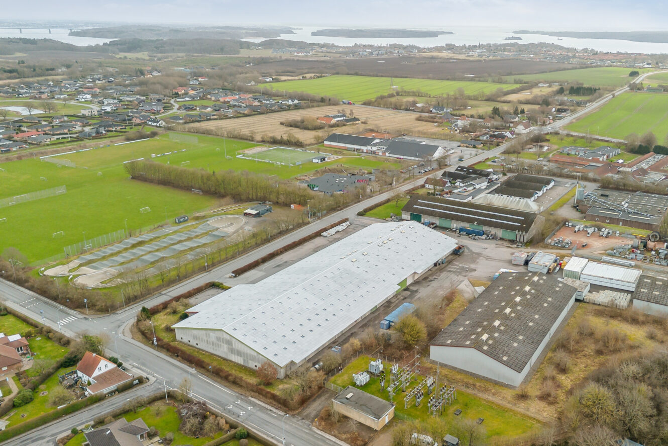 Luftfoto af et industriområde med lagerbygninger, sportspladser, boliger, veje, landbrugsjord og vand med øer i det fjerne.