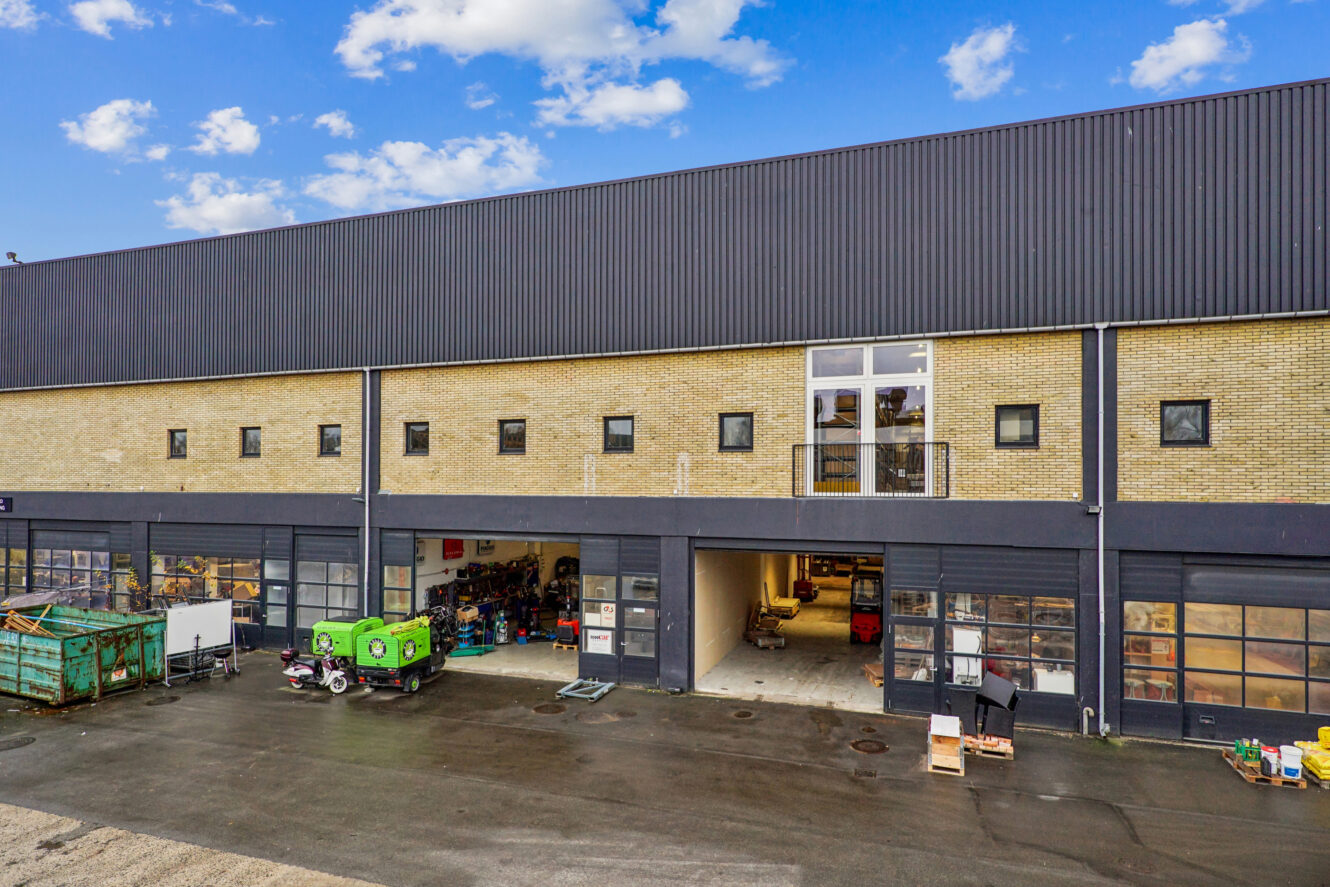 En stor industribygning med flere garageporte, en murstensfacade, små vinduer samt udstyr og containere på fortovet udenfor.