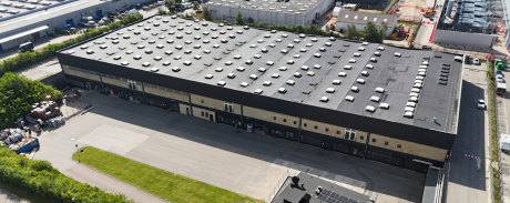 Luftfoto af et stort industrilager med fladt tag, omgivet af veje og nabobygninger i et industriområde.
