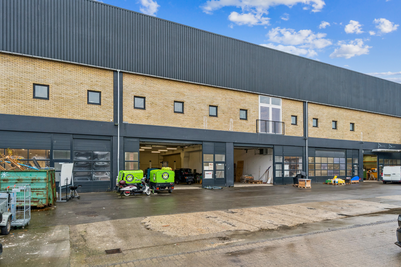 Stor industribygning med åbne garageporte, udstyr og køretøjer udenfor, under en delvis overskyet himmel.