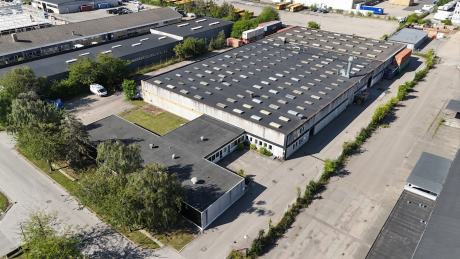 Luftfoto af et industrielt lagerkompleks bestående af en stor bygning med fladt tag, omgivet af veje, træer og nabobygninger.