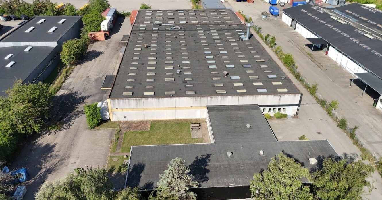 Luftfoto af en stor industribygning med fladt tag, adskillige ovenlys, omgivende træer og tilstødende befæstede arealer.