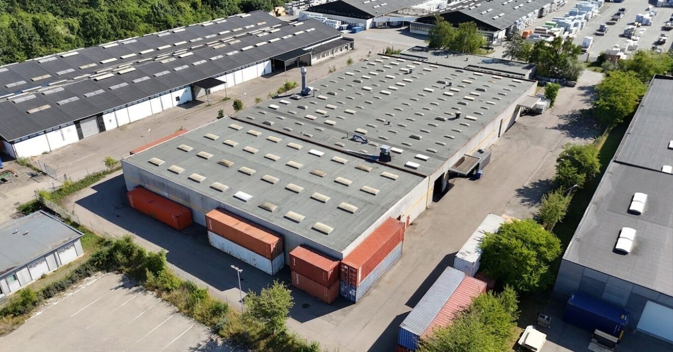 Luftfoto af et industrilager med fladt tag, omgivet af fragtcontainere, parkeringsarealer og nærliggende bygninger.