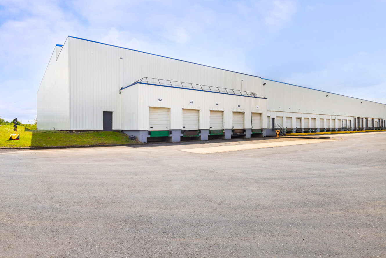 Grand entrepôt industriel, plusieurs portes de quai de chargement blanches, une rampe et une large zone asphaltée sous un ciel partiellement nuageux.