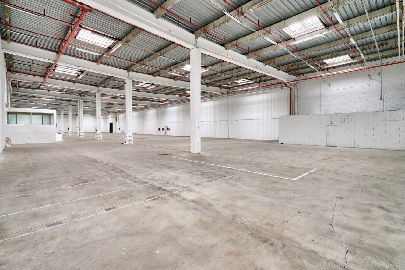 Spacieux entrepôt industriel vide avec sols en béton, murs blancs, hauts plafonds et poutres métalliques apparentes. Un petit bureau fermé est visible à l'arrière-plan.
