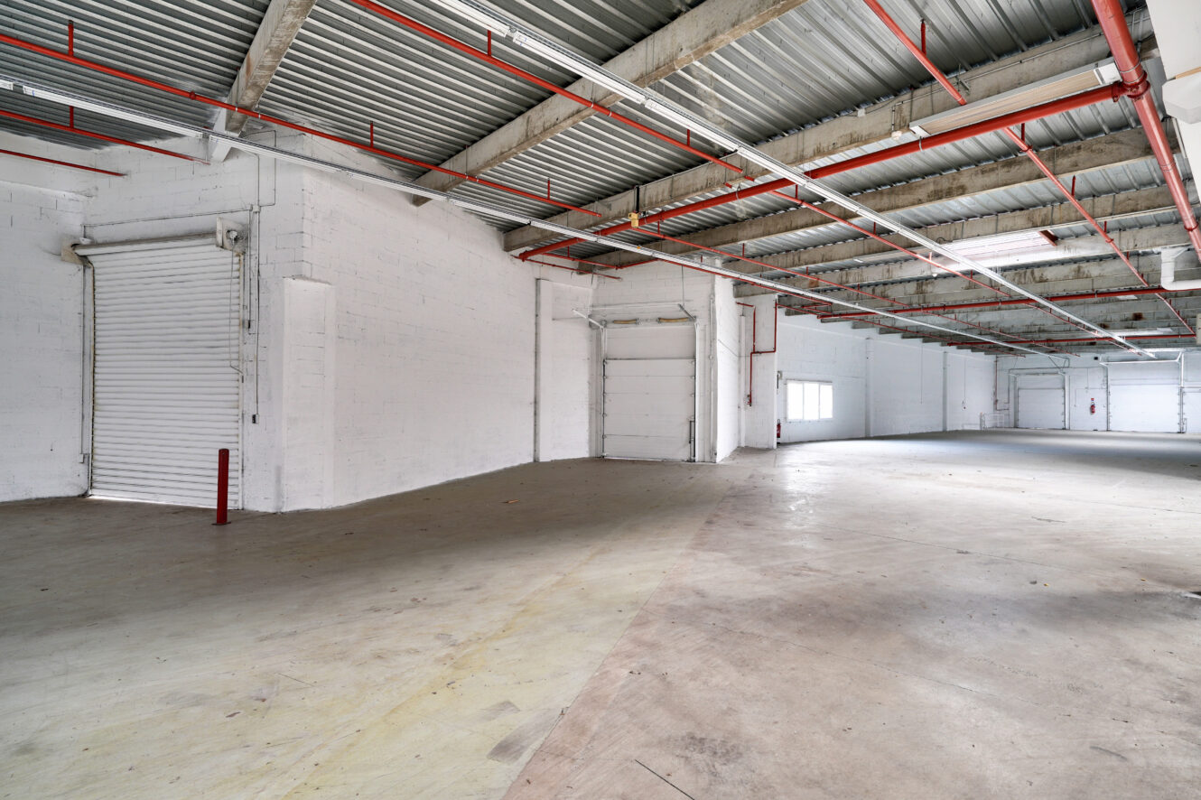 Intérieur d'un entrepôt industriel vide avec plafond apparent, murs en briques peintes en blanc, portes roulantes et sol en béton.