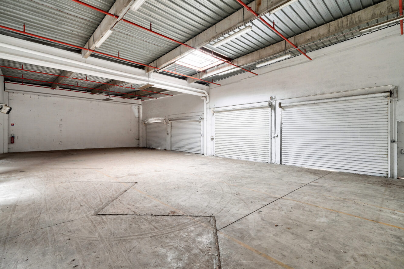 Intérieur d'un entrepôt vide avec un sol en béton, des murs blancs, un plafond métallique, des plafonniers, trois grandes portes roulantes et un extincteur visible sur le mur de gauche.