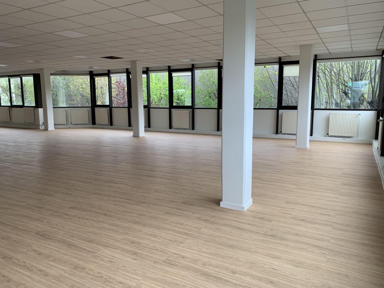 Grande pièce vide avec un parquet en bois clair, des murs blancs, des colonnes de soutien et de nombreuses fenêtres laissant entrer la lumière naturelle.