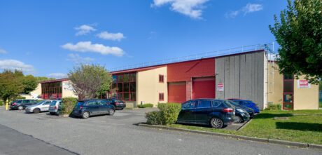 Un entrepôt commercial aux accents rouges et aux grandes fenêtres, plusieurs voitures garées devant, des arbres et un ciel bleu clair avec des nuages épars.