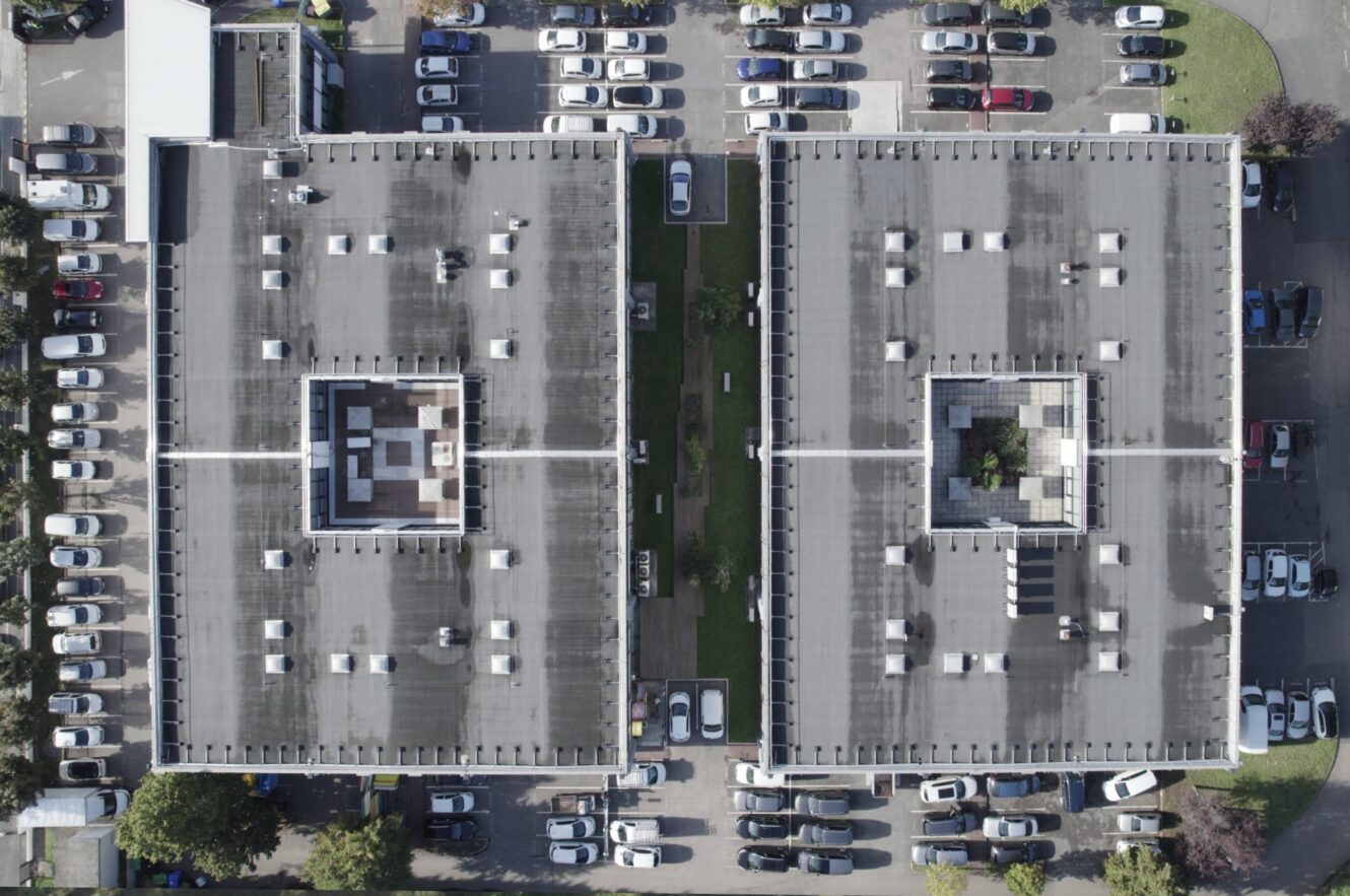 Vue aérienne de deux bâtiments rectangulaires à toit plat, séparés par une bande verte, entourés de voitures garées dans un parking.