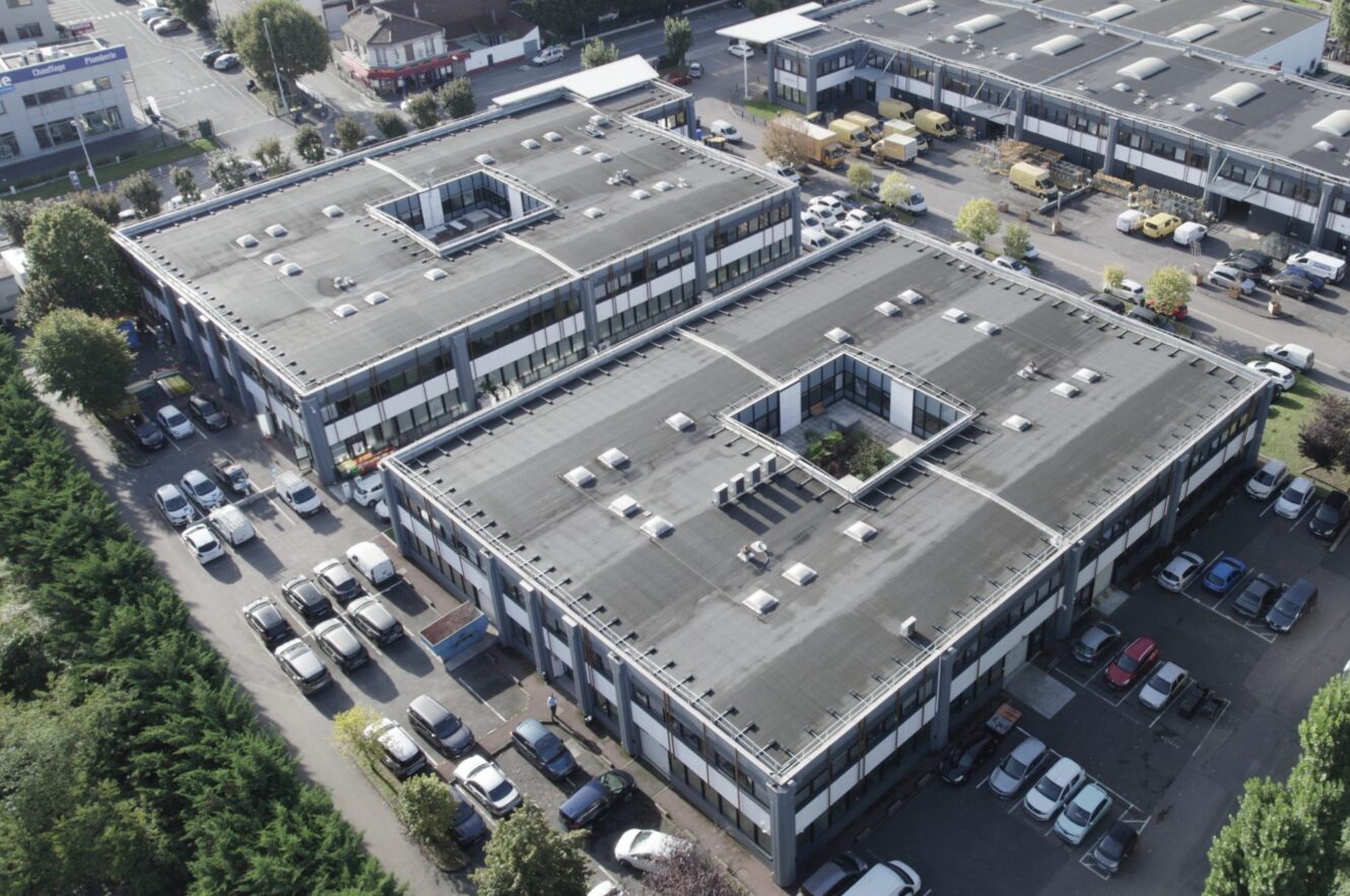 Vue aérienne de deux grands immeubles de bureaux rectangulaires avec cour centrale, entourés de voitures garées et de routes adjacentes.