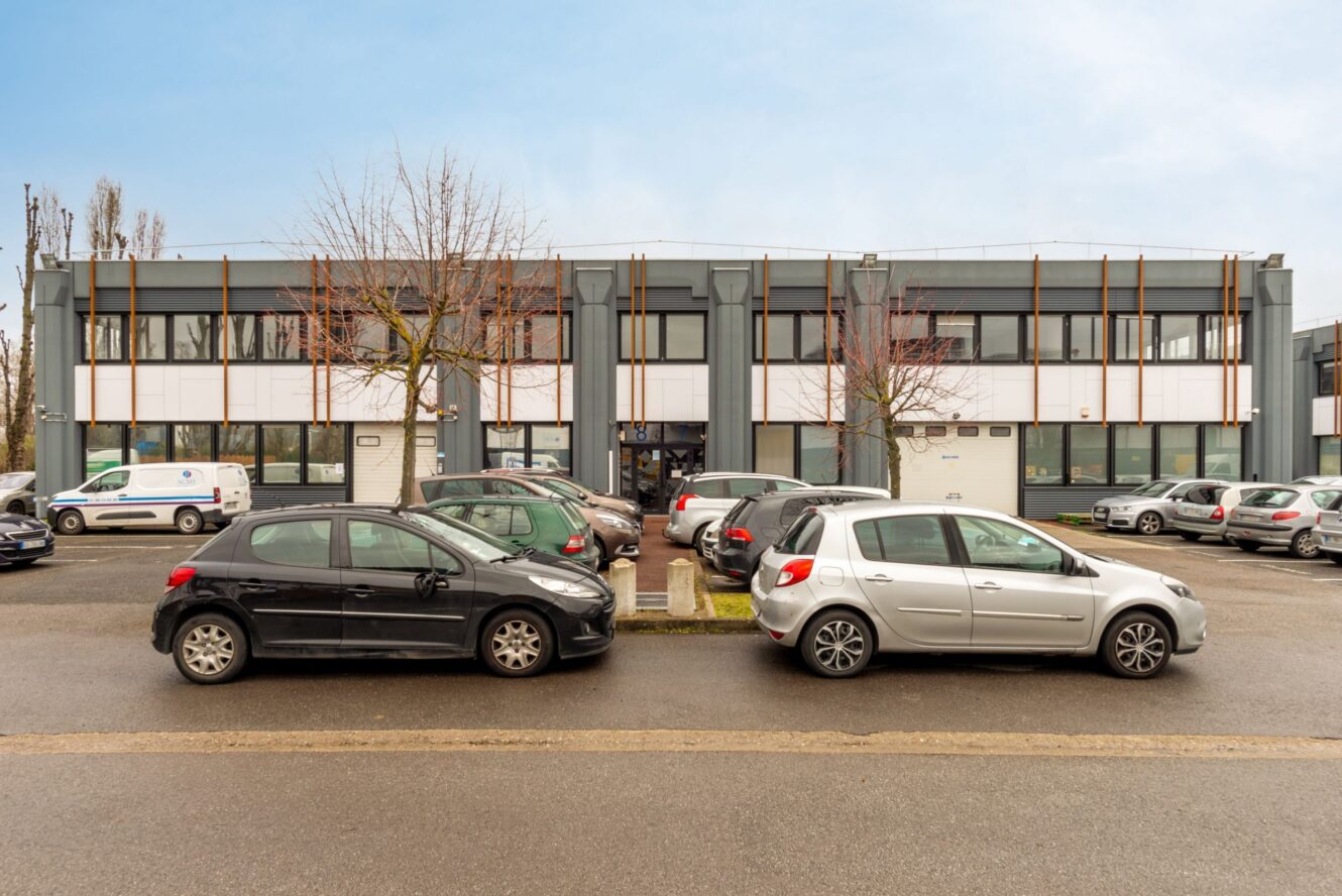 Un bâtiment commercial de deux étages avec de grandes fenêtres et un parking à l'avant, où plusieurs voitures sont garées par un temps couvert.