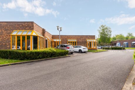Un immeuble de bureaux en briques avec des fenêtres jaunes, plusieurs voitures garées, des haies taillées et un parking asphalté vide sous un ciel partiellement nuageux.