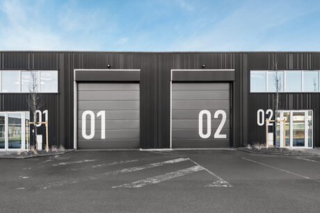 Un bâtiment industriel moderne avec deux grandes portes de garage grises étiquetées 01 et 02, flanquées de portes d'entrée en verre portant les mêmes numéros.
