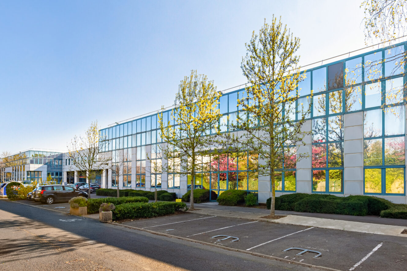 Immeuble de bureaux moderne avec de grandes baies vitrées, entouré d'arbres et de verdure, avec des voitures garées dans un parking sous un ciel bleu clair.