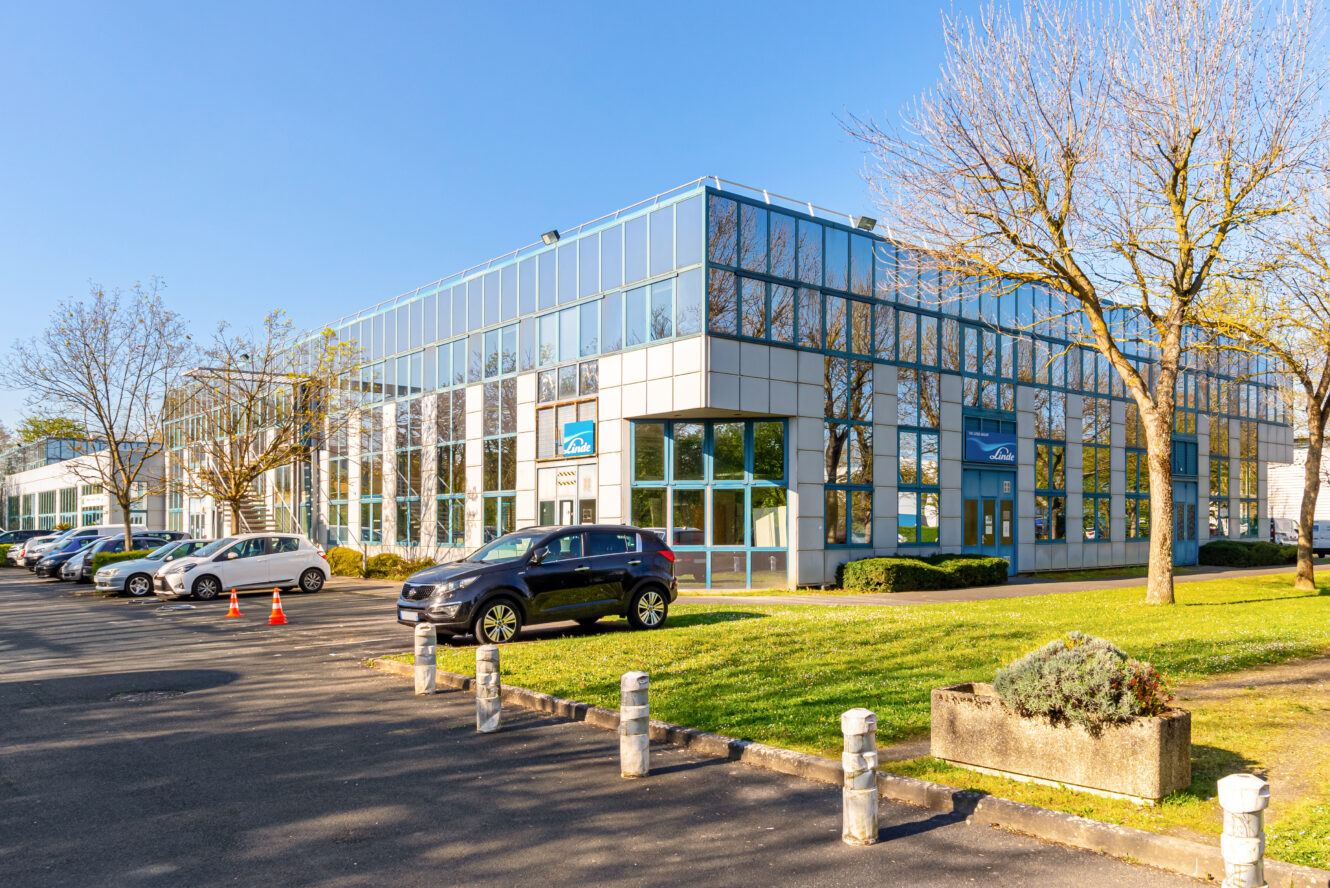 Un immeuble de bureaux moderne en verre avec des voitures garées devant, entouré de pelouses et d'arbres verts par une journée claire et ensoleillée.