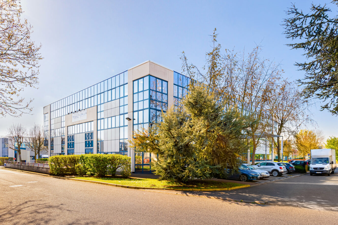 Immeuble de bureaux moderne en verre avec des fenêtres réfléchissantes, entouré d'arbres, d'arbustes et d'un parking avec plusieurs voitures par une journée claire et ensoleillée.