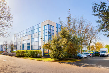 Immeuble de bureaux moderne en verre avec des fenêtres réfléchissantes, entouré d'arbres, d'arbustes et d'un parking avec plusieurs voitures par une journée claire et ensoleillée.