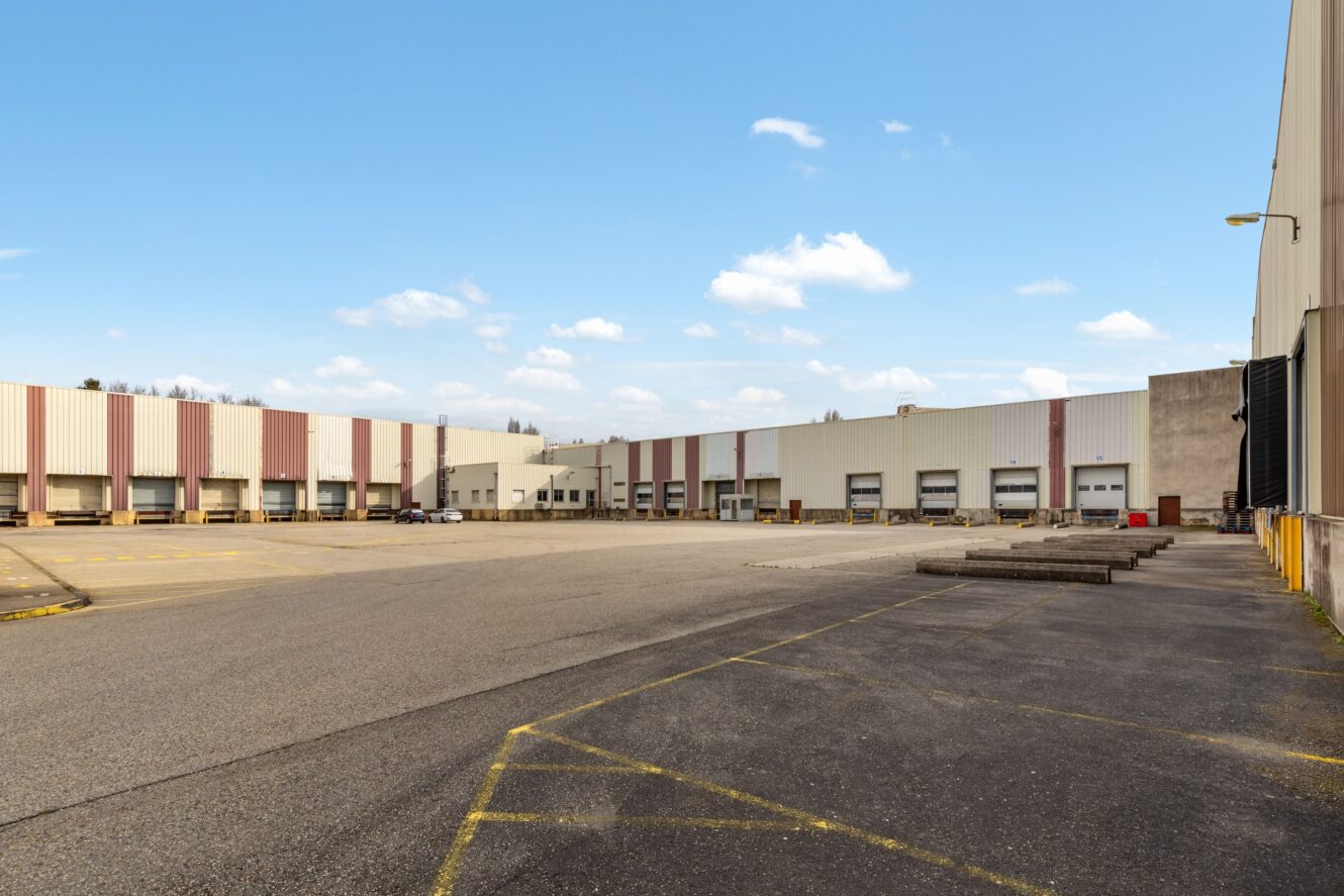 Grand bâtiment d'entrepôt avec plusieurs quais de chargement et un vaste parking asphalté vide sous un ciel partiellement nuageux.