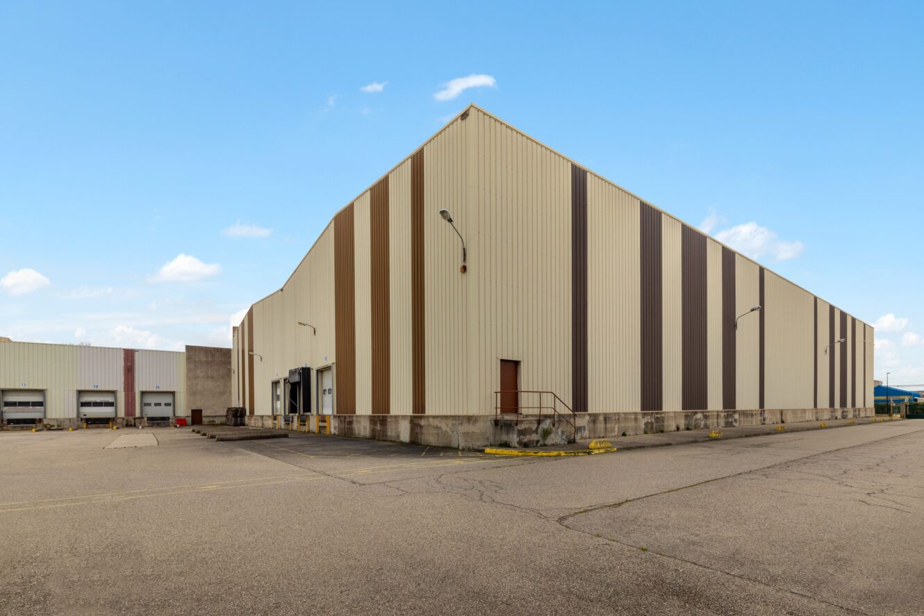 Un grand entrepôt industriel avec des rayures verticales marron et beige, des quais de chargement, un petit escalier métallique et un terrain asphalté en grande partie vide sous un ciel bleu.