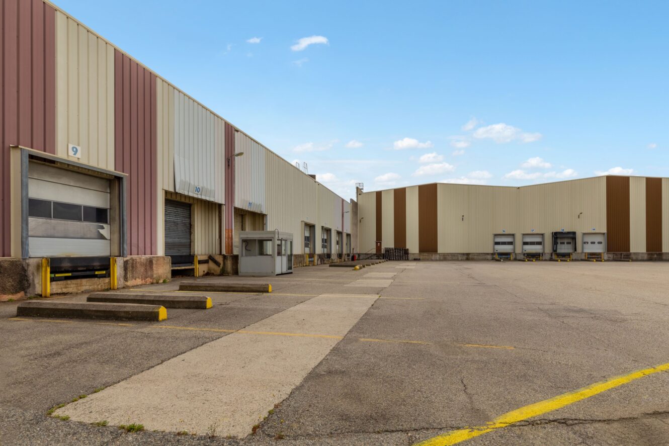 Grand entrepôt industriel avec plusieurs quais de chargement et portes basculantes, entouré d'un terrain pavé vide sous un ciel généralement dégagé.