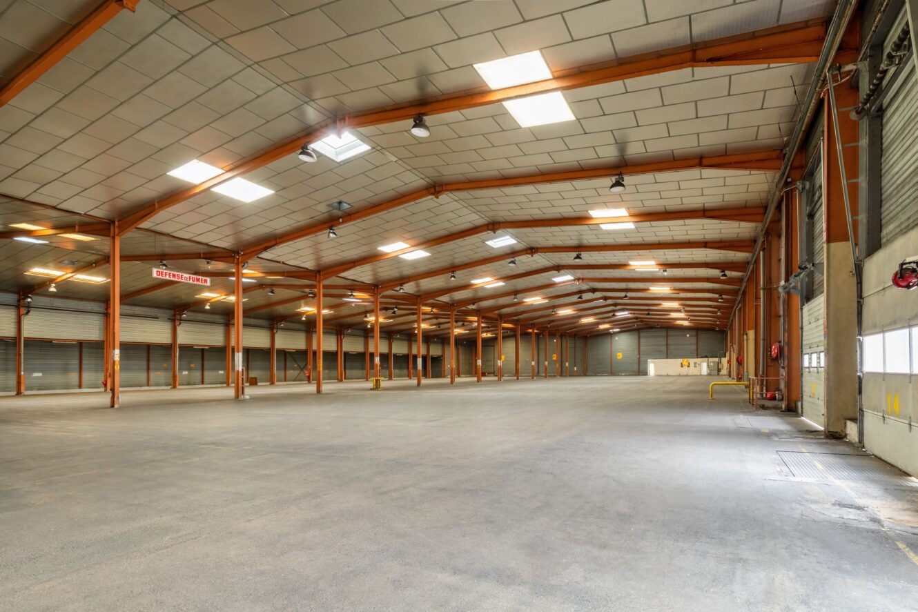 Intérieur d'un entrepôt vide et spacieux avec un haut plafond, des poutres métalliques apparentes et de grands plafonniers. Un sol en béton et plusieurs portes de quai de chargement sont visibles sur le côté droit.