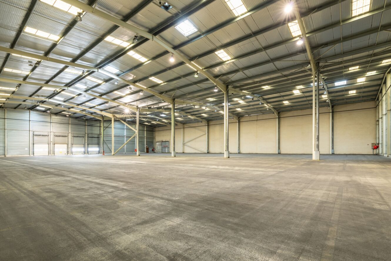 Intérieur d'un grand entrepôt vide avec de hauts plafonds, des sols en béton, des poutres de soutien en métal et un éclairage zénithal intense.