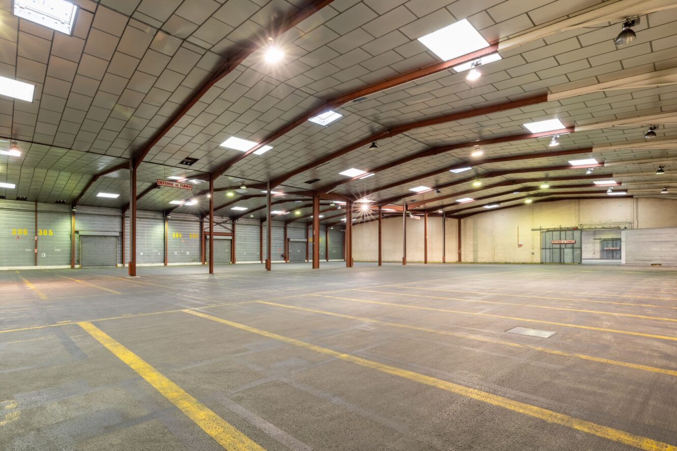 Intérieur d'un entrepôt vide avec de hauts plafonds, des poutres de soutien métalliques, un sol en béton et des lignes de stationnement marquées. Des lampes fluorescentes sont allumées et de grandes portes roulantes bordent le mur du fond.