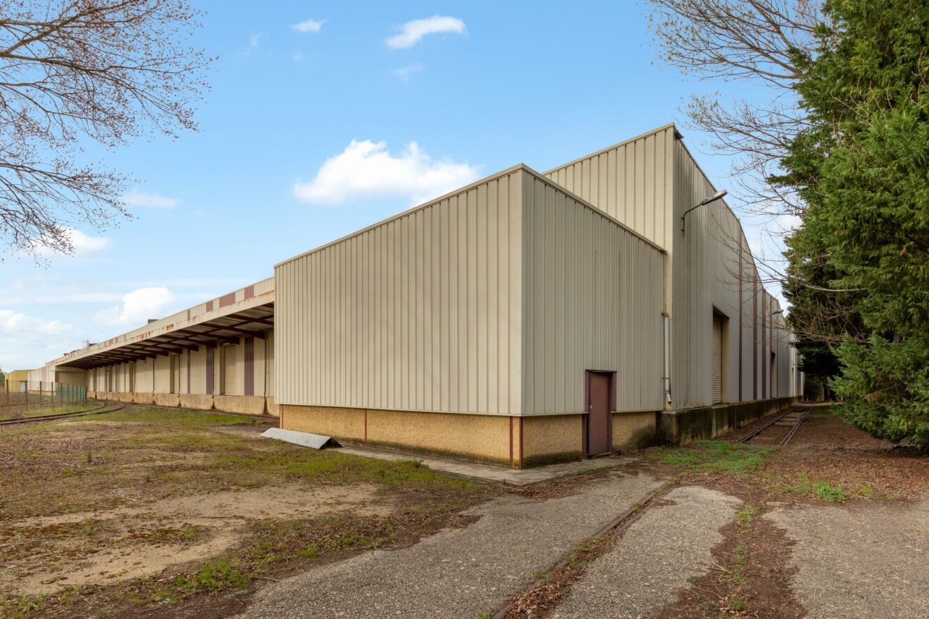 Un grand entrepôt industriel beige avec un extérieur métallique et une porte marron, entouré d'une végétation et d'arbres clairsemés, sous un ciel dégagé.