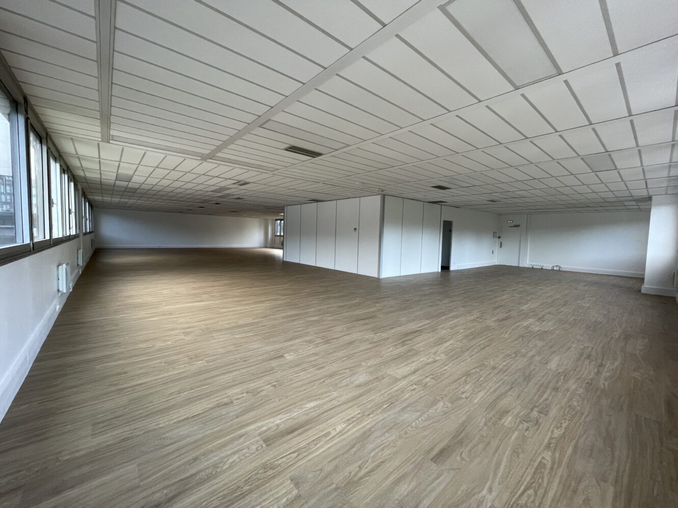 Grand espace de bureau vide avec un sol en bois clair, des murs blancs, un plafond quadrillé et des fenêtres sur un côté. Des pièces cloisonnées sont visibles vers l'arrière.