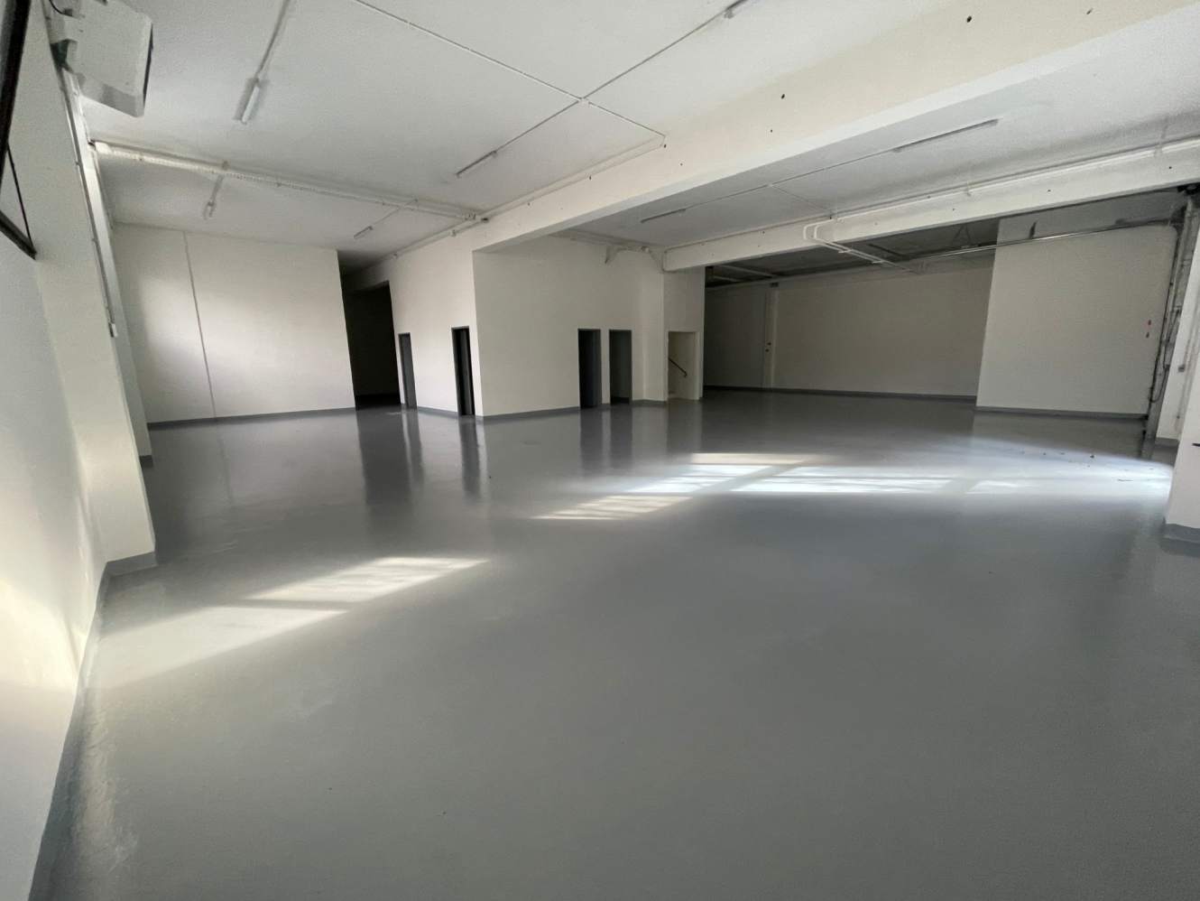 Espace industriel vide avec des sols gris lisses, des murs blancs, un haut plafond et plusieurs portes ; la lumière naturelle pénètre par les fenêtres.